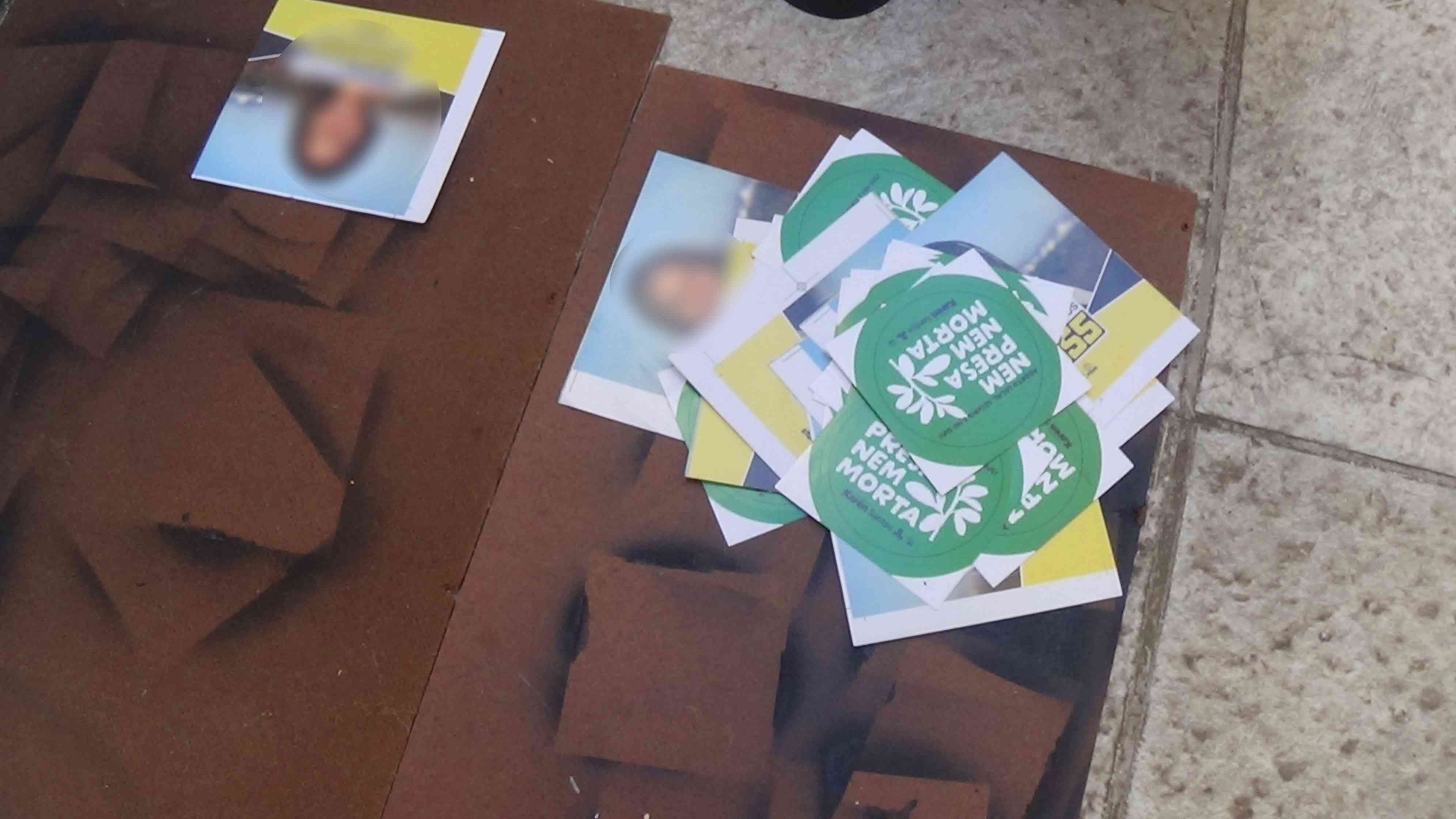 Foto de vários adesivos da época eleitoral do passado sobre uma tábua de madeira que está no chão.