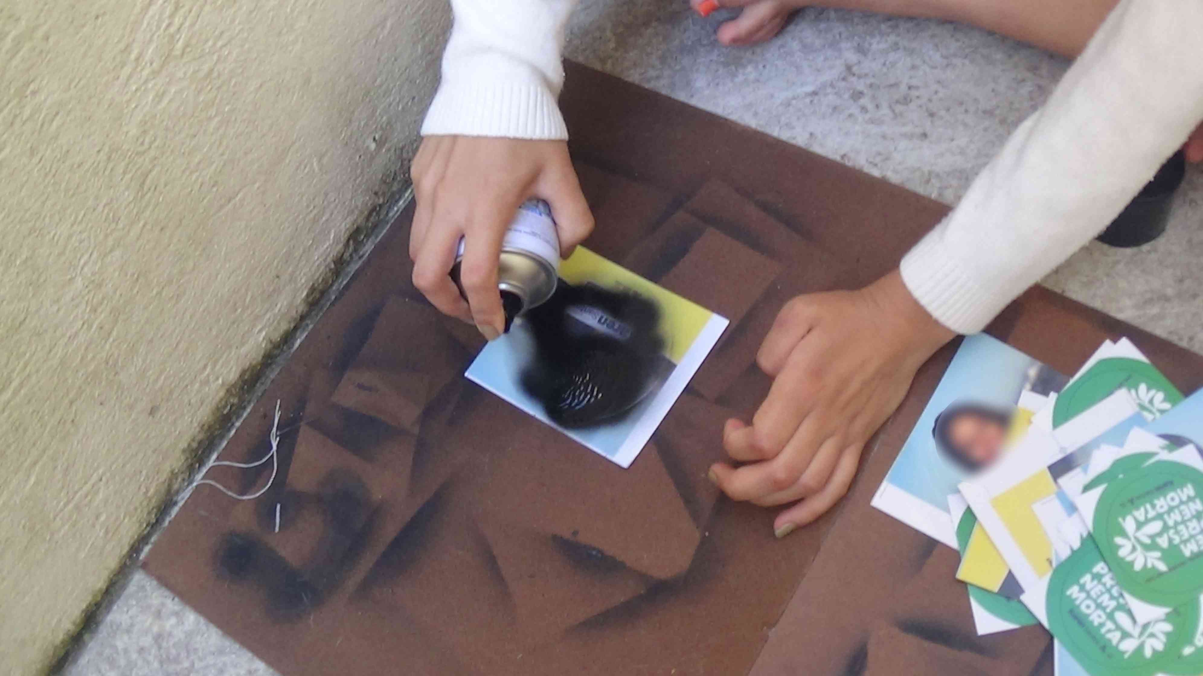 Foto de pessoa sentada no chão passando spray preto em cima do adesivo que está sobre a tábua de madeira no chão