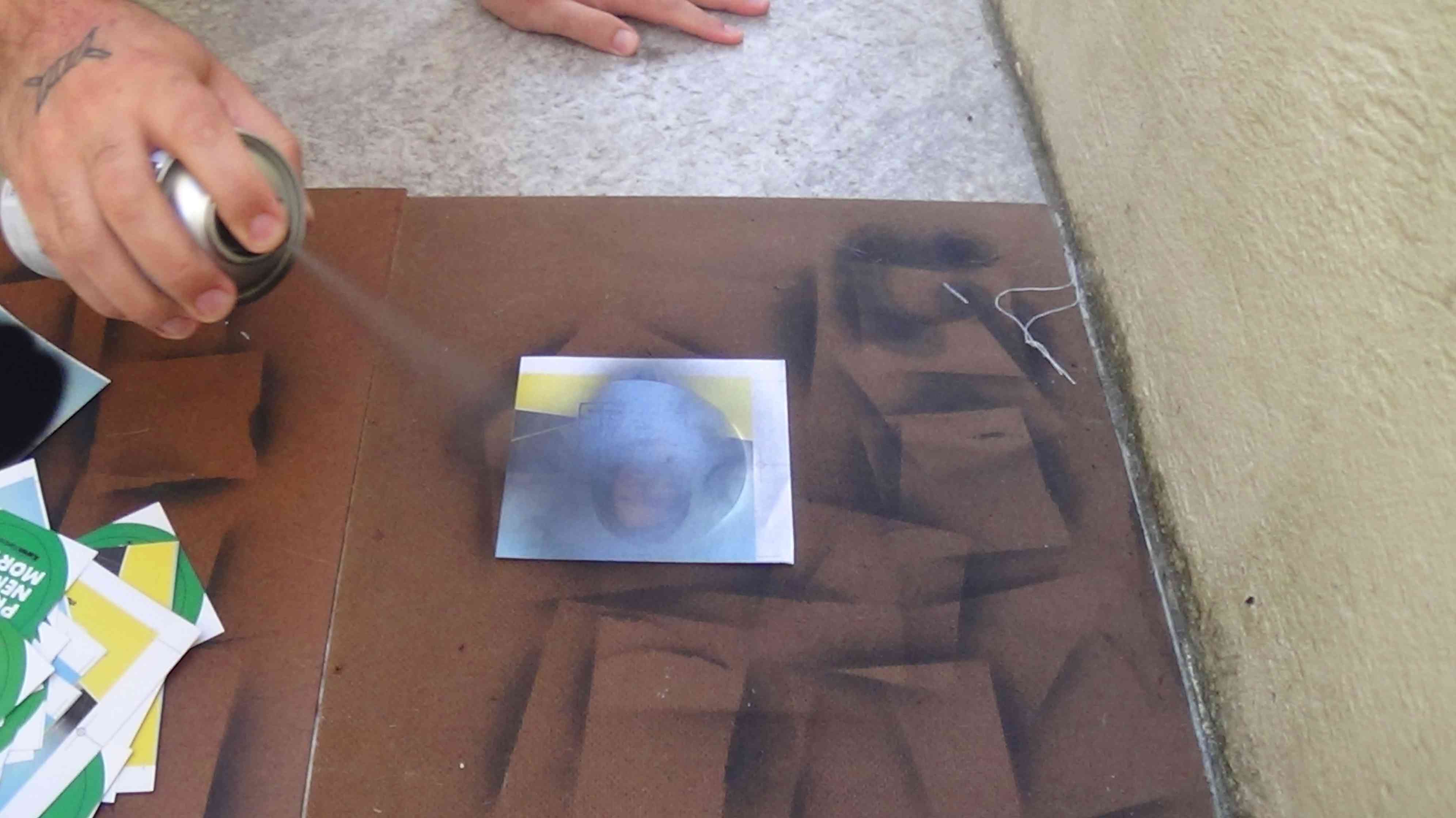 Foto uma mão segurando um spray sendo apertado, é possível ver a tinta saindo e colorindo um adesivo que antes tinha um conteúdo e agora está ficando todo preto, em cima de tábua de madeira.