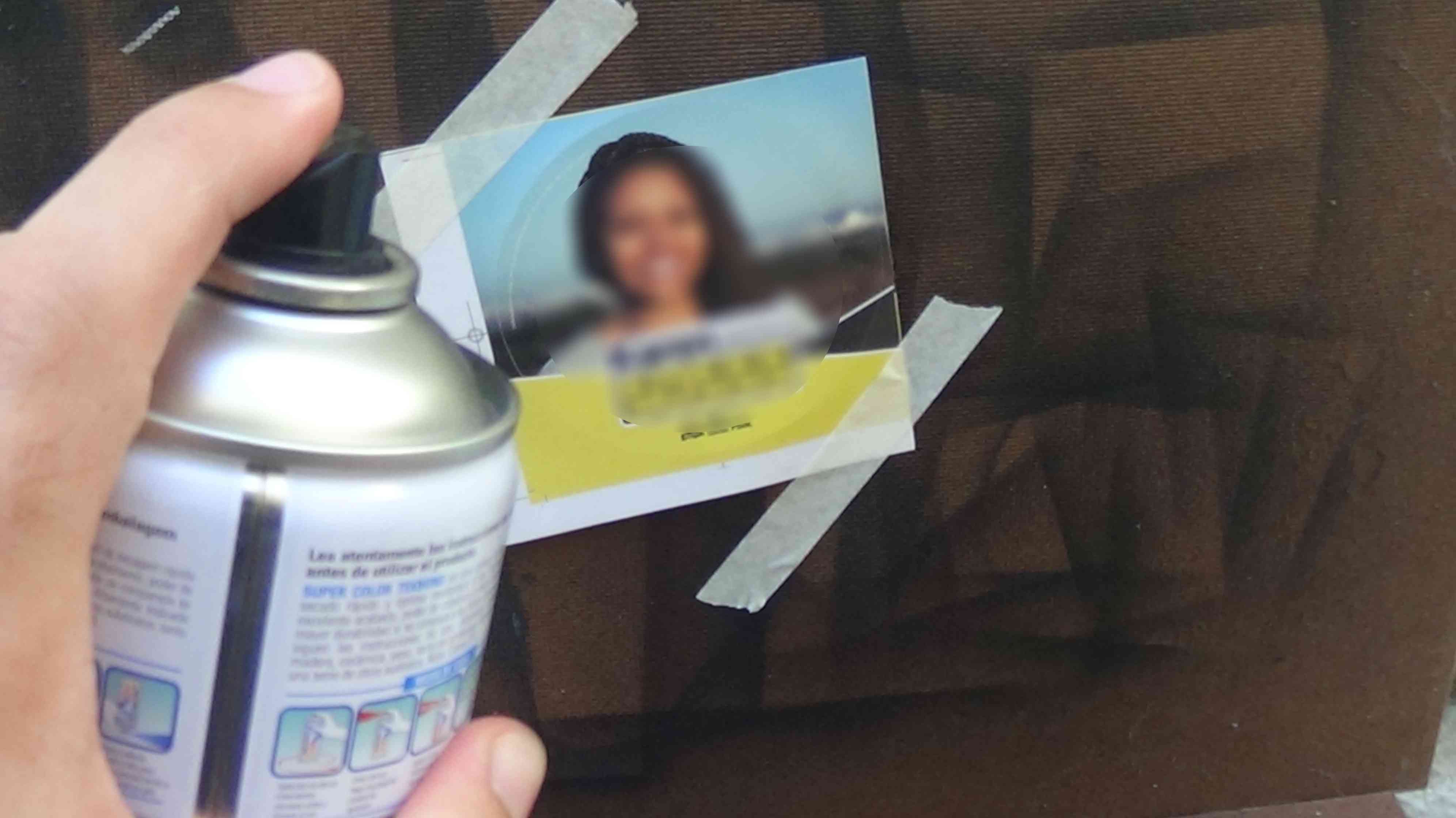 Foto de uma mão segurando spray em direção a um adesivo eleitoral que está colado em uma tábua de madeira que está encostada na parede. O adesivo está grudado com duas fitas crepes.