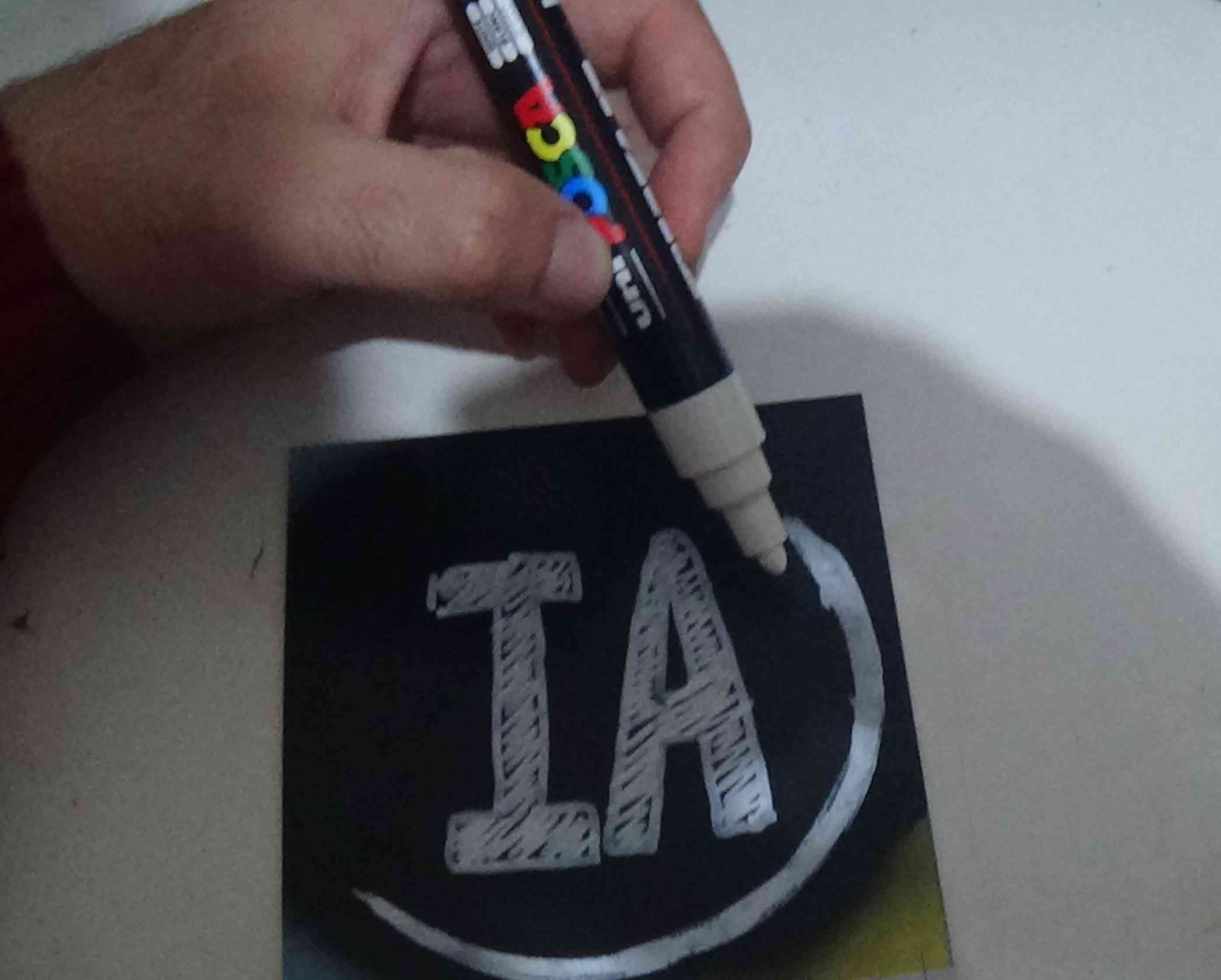 Foto de um adesivo preto sendo preenchido com tinta branca de uma caneta poska. É possível ver as letras IA e um círculo sendo traçado ao seu redor, enquanto uma mão segura a caneta poska e finaliza o círculo.