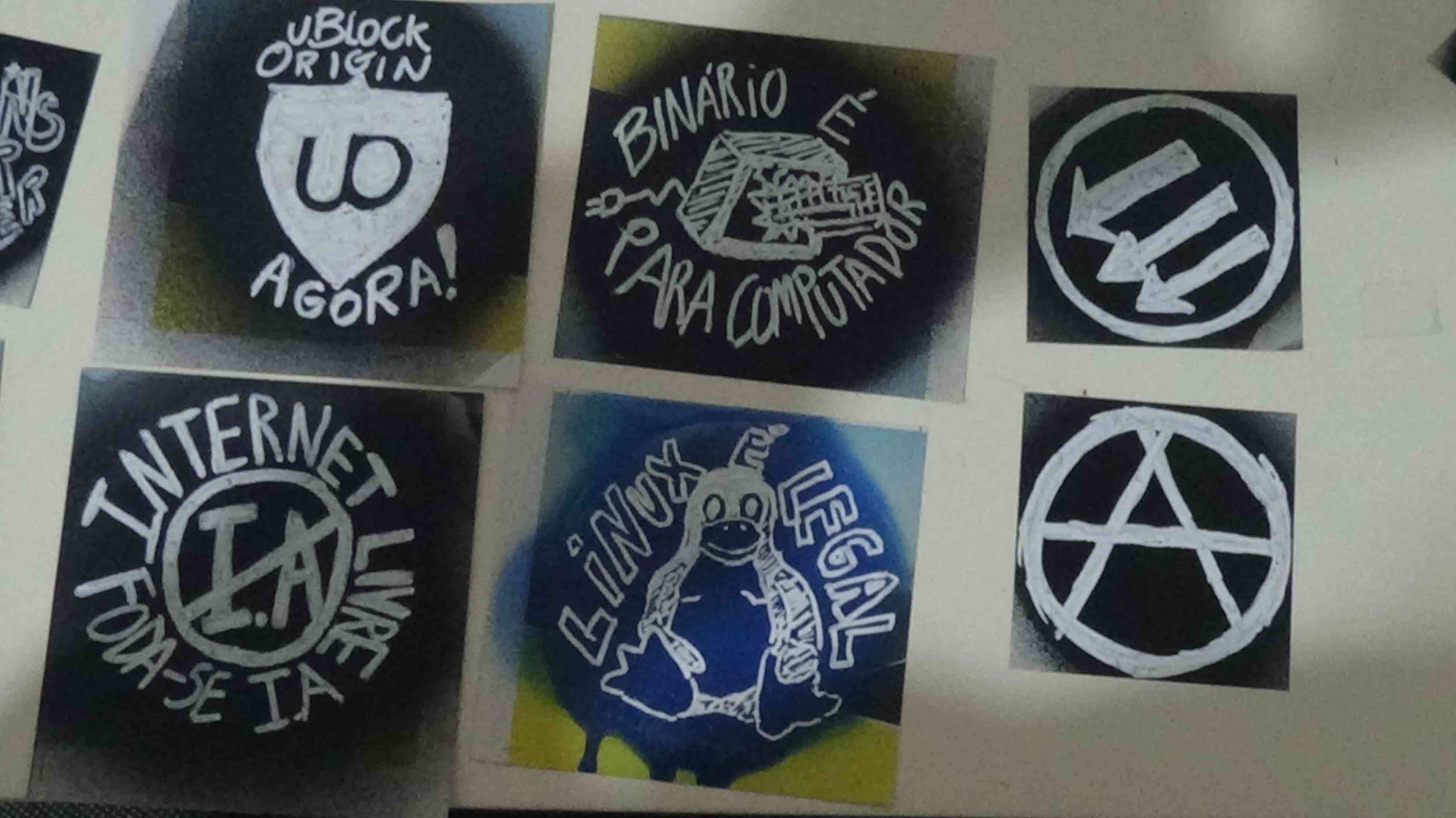 Foto de 6 adesivos prontos: 1 uBlock Origin Agora!, 2 Binário é para computador e um teclado quebrando uma tela de PC, 3 Símbolo das três setas apontadas para a diagonal esquerda, 4 Internet Livre Foda-se IA, 5 Linux é Legal com o Tux no meio, 6 A na bola.