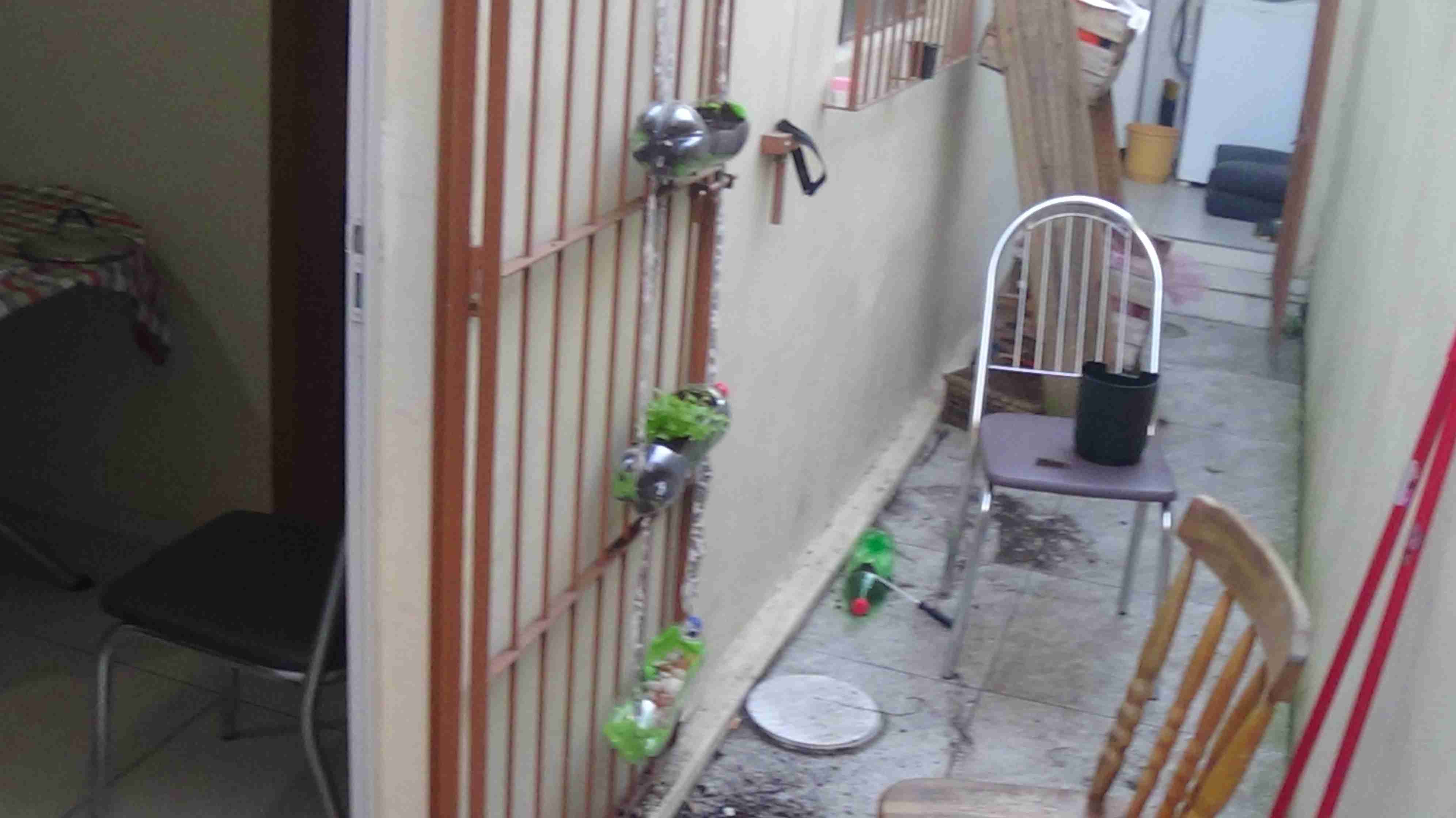 Parte externa de uma casa onde é possível ver uma horta vertical de garrafas pet sendo construída. A garrafa de cima está coberta de terra, a do meio tem uma planta em seu centro, e a de baixo está sendo construída, com pedras e o recorte da ecobag.