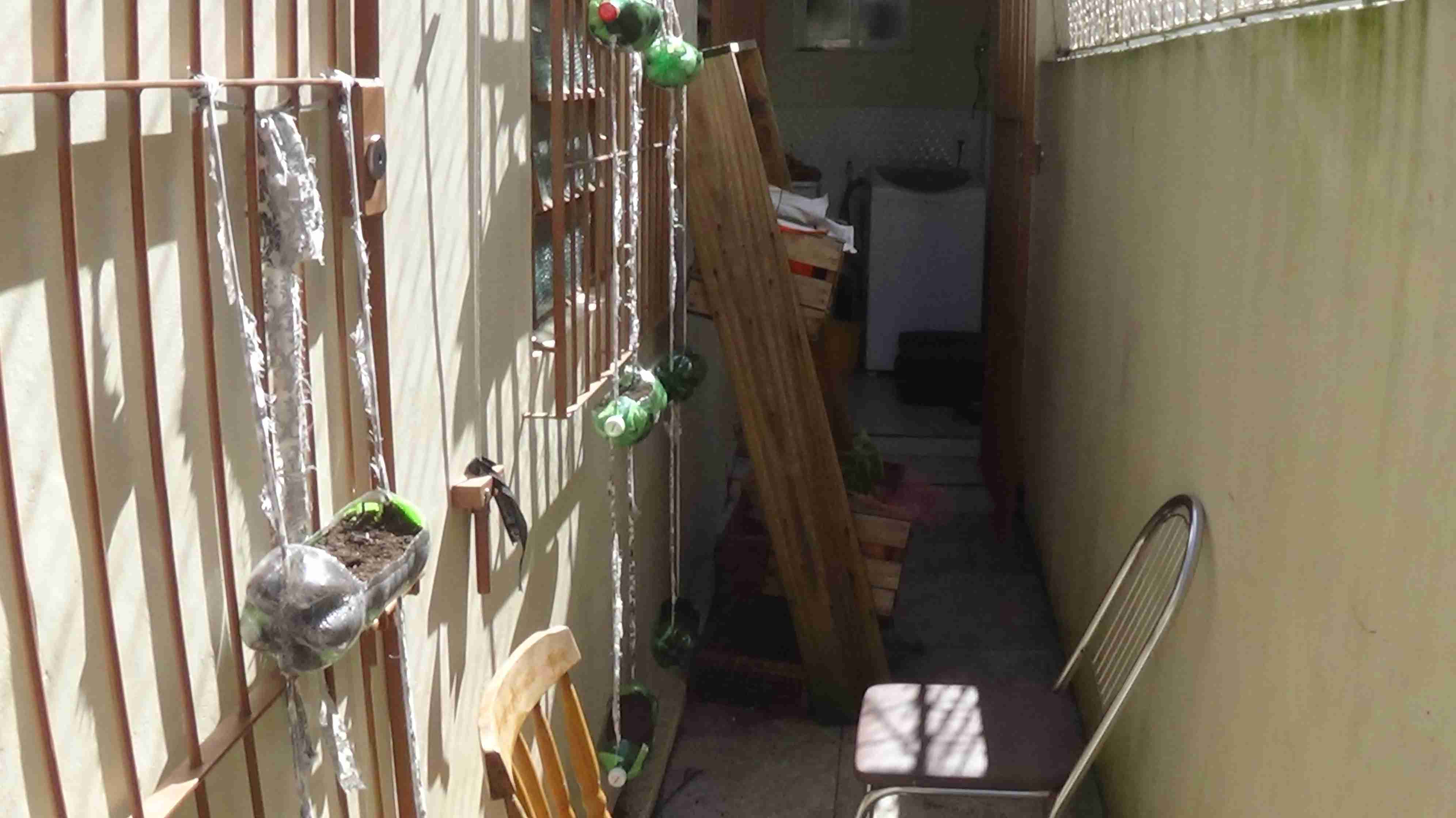 Parte externa de uma casa sob o sol. É possível ver as 3 hortas verticais uma ao lado do outra e penduradas nas grades. Todas as garrafas pets na horizontal que aparecem na foto estão cobertas com terra.