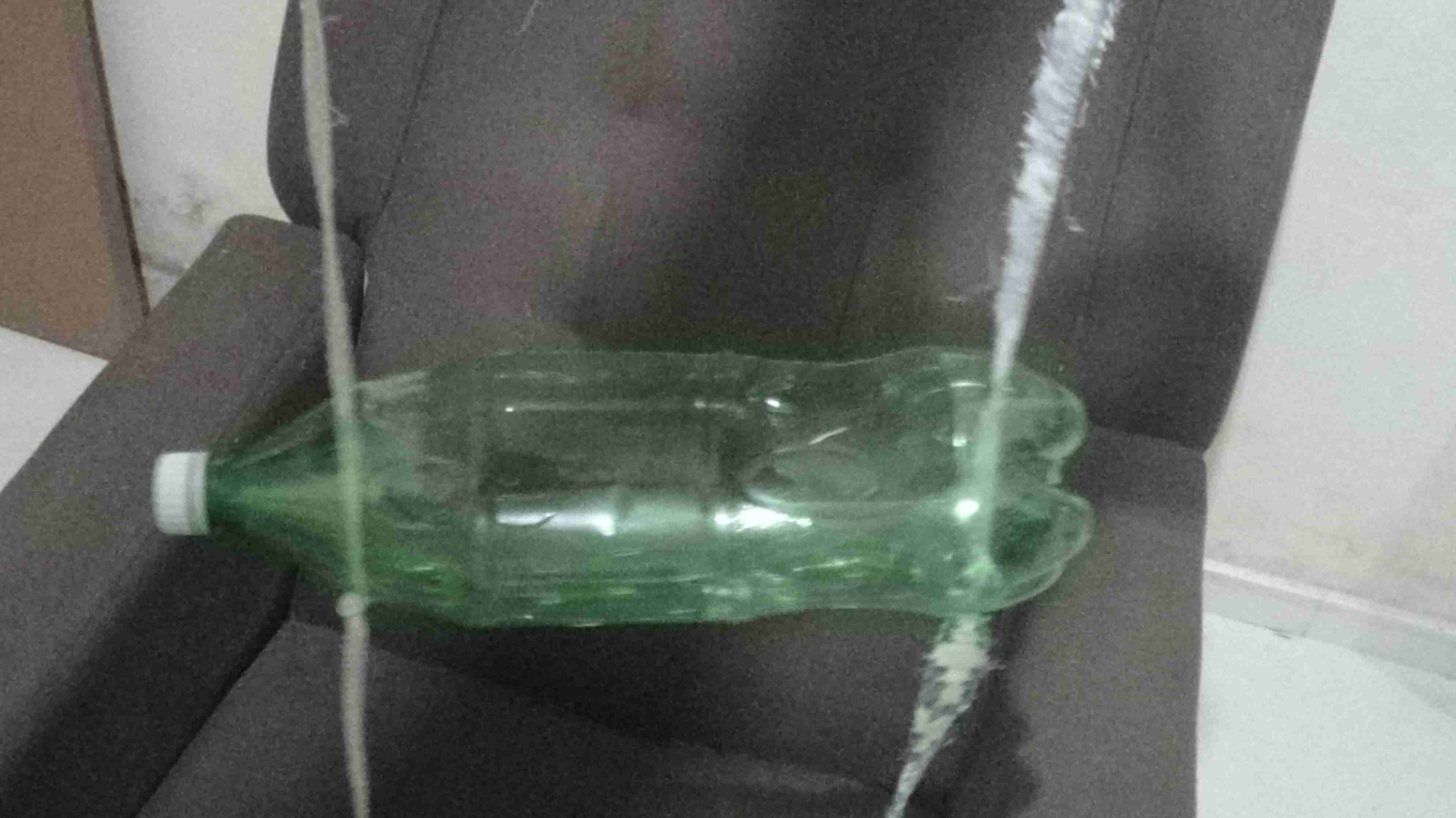 Garrafa pet na horizontal suspensa no ar acima de uma poltrona, é possível ver os 2 fios atravessando-a e sustentando-a à partir de um nó em cada fio.